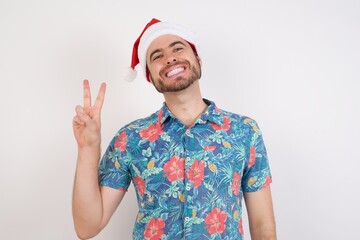 Young caucasian man wearing hawaiian shirt and Santa hat over isolated white background smiling with happy face winking at the camera doing victory sign. Number two.