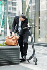 Stylidh young entrepreneur with scooter searching for document in leather bag when talking on phone with colleague