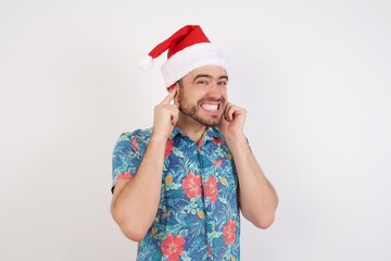 Young caucasian man wearing hawaiian shirt and Santa hat over isolated white background covering ears with fingers with annoyed expression for the noise of loud music. Deaf concept.