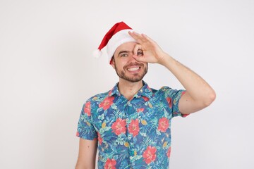 Young caucasian man wearing hawaiian shirt and Santa hat over isolated white background doing ok gesture with hand smiling, eye looking through fingers with happy face.