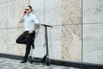 Confident stylish young entrepreneur leaning on wall next to his scooter and calling on phone