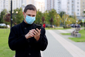 Coronavirus protection, man in medical mask walking on a autumn city street with smartphone in hands. Concept of quarantine during covid-19 pandemic