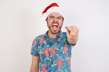 Young caucasian man wearing hawaiian shirt and Santa hat over isolated white background pointing displeased and frustrated to the camera, angry and furious ready to fight with you.