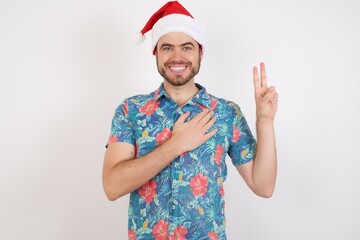 Young caucasian man wearing hawaiian shirt and Santa hat over isolated white background smiling swearing with hand on chest and fingers up, making a loyalty promise oath.