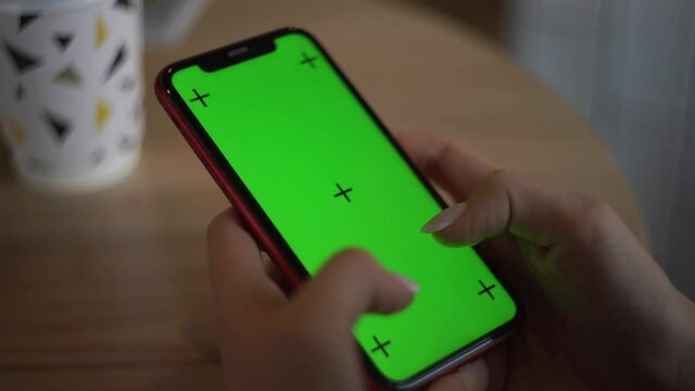 Girl in a cafe sits at beige table with white cardboard craft coffee. Hold your phone with green screen for insertion and tracking. Scrolling and swiping smartphone screen