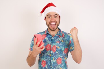 Positive Young caucasian man wearing hawaiian shirt and Santa hat over isolated white background holds modern cell phone connected to headphones, clenches fist from good emotions, exclaims with joy,