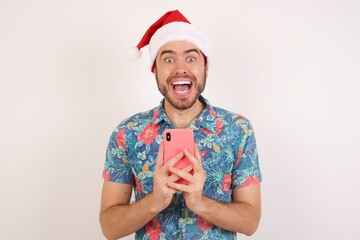 Copyspace photo of cheerful Young caucasian man wearing hawaiian shirt and Santa hat over isolated white background holding phone in his hands while