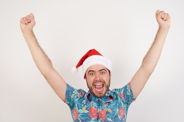 Photo of shouting screaming Young caucasian man wearing hawaiian shirt and Santa hat over isolated white background having won contests seeing sales discounted goods with hands up amazed