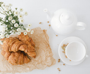 Top view of white cup and teapot with tasty croissants on bright table.