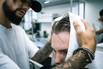 Fototapeta premium Tattooed barber with towel drying hair of client before haircut in salon 