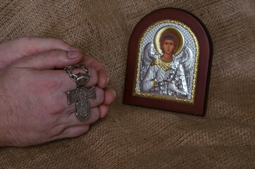 Hands folded in prayer with a silver cross in front of the icon of the Archang