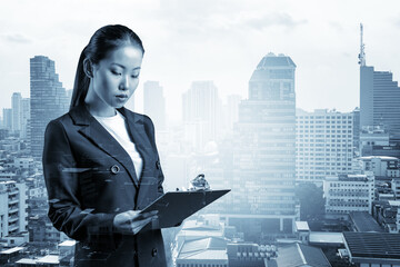 Attractive young Asian business woman in suit taking notes into the notebook, new career opportunities, MBA assignment. Bangkok on background. Double exposure.