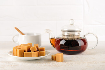 Pile of caramel vanilla fudge on a white ceramic saucer in front of white cup and transparent glass tea pot over white wood table. Fresh tasty candies made of milk and sugar. Tea drinking with sweets.