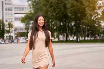 Asian girl in a beautiful tight-fitting beige dress walks through the city streets and looks away.