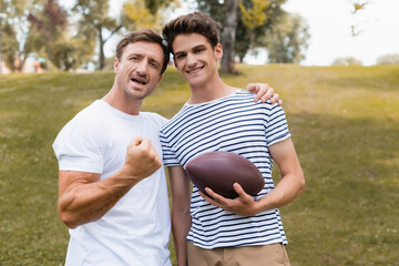 excited father showing clenched fist and standing near teenager son with rugby ball