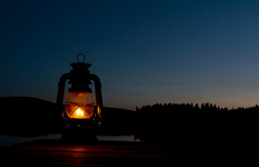 Old kerosene lantern with warm yellow light on a bridge by a lake in the evening.