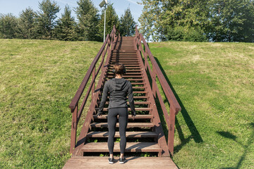 Fit caucasian woman stands in front of wooden staircase in city park ready for outdoor exercise. Rear view. Sport background. Overcoming obstacles theme.