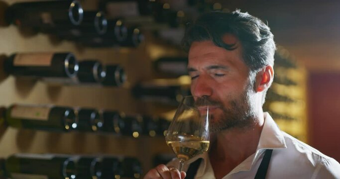 Authentic shot of successful male sommelier is tasting a flavor and checking white wine quality poured in transparent glass in a wine cellar.