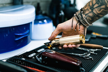 Cropped view of young tattooed barber holding hair clipper near equipment on table in barbershop 