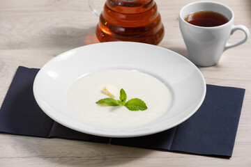 Semolina milk porridge in a white plate with butter and mint leaves on a wooden background with a mug of tea. Healthy Breakfast, proper morning.