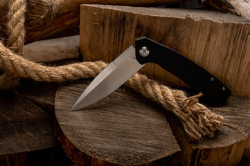 Knife with a black handle in a bent position. A folding knife on a tree stump with a rope.