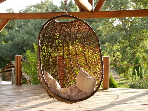 A wicker chair with cushions is suspended from the strong wooden beams of the veranda against the background of green trees on a Sunny summer day . A place to relax in the country style.