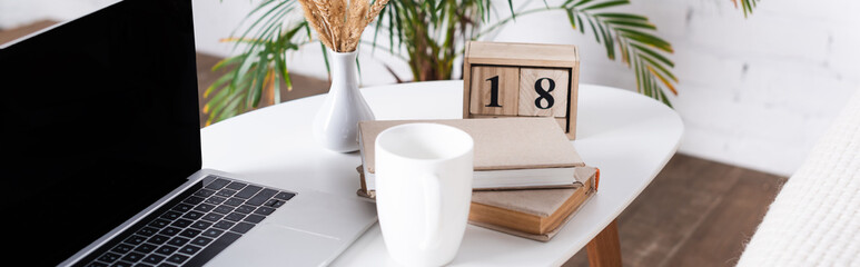 Panoramic shot of laptop with blank screen, books and cup on coffee table