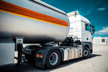 Truck with trailer, tank with flammable liquid, sunny day outside, metallic color container, blue sky with white clouds, gravel