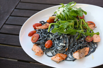Black spaghetti pasta colored with squid ink, cream sauce, salmon, tomatoes and rocket salad in a white plate on a dark rustic wooden garden table, selected focus