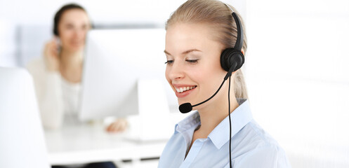 Blond woman call operator is using computer and headset for consulting clients online. Group of...