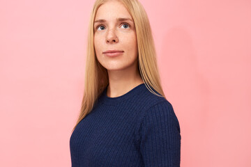 Portrait of gorgeous beautiful young blonde Caucasian female with freckles and blue eyes posing isolated in dark blue dress, looking up with thoughtful dreamy facial expression, making future plans