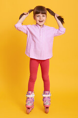 Little girl roller skating against yellow background, wearing shirt and leggins, touching her hair,...