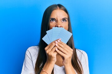 Young hispanic woman playing gambling poker covering face with cards angry and mad screaming frustrated and furious, shouting with anger looking up.