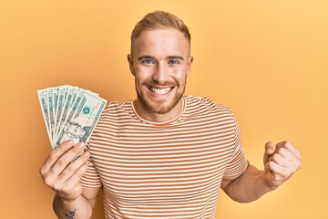 Young caucasian man holding dollars screaming proud, celebrating victory and success very excited with raised arm