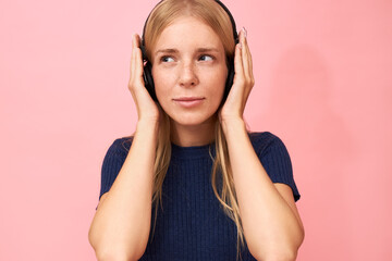 People, technology, leisure and fun concept. Studio image of cute young female with freckles and facial piercing listening nice music via online streaming service, having joyful pleased look