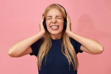 Isolated image of funny emotional young European female with teeth braces wearing wirless headphones closing eyes, holding hands on ears, enjoying loud high quality music against pink background