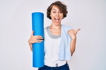 Young hispanic woman holding yoga mat pointing thumb up to the side smiling happy with open mouth