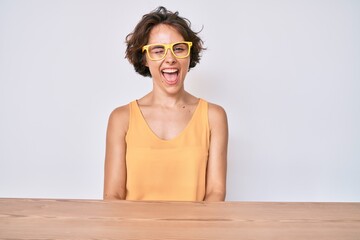 Young hispanic woman wearing casual clothes and glasses sitting on the table winking looking at the...