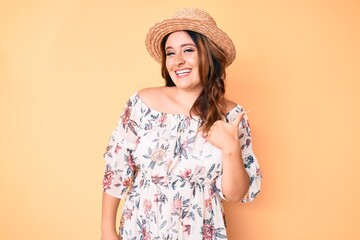 Young beautiful caucasian woman wearing summer dress and hat doing happy thumbs up gesture with hand. approving expression looking at the camera showing success.