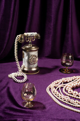 Vintage style concept with telephone, crystal glass and pearl necklace on violet curtain...