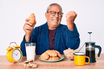 Senior handsome man with gray hair sitting on the table eating croissant for breakfast annoyed and frustrated shouting with anger, yelling crazy with anger and hand raised