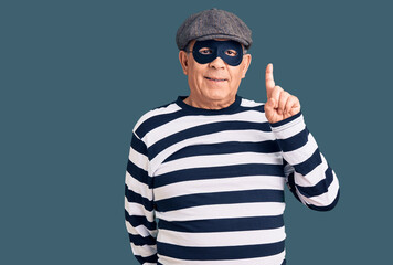 Senior handsome man wearing burglar mask and t-shirt showing and pointing up with finger number one while smiling confident and happy.