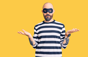 Young handsome man wearing burglar mask clueless and confused with open arms, no idea concept.