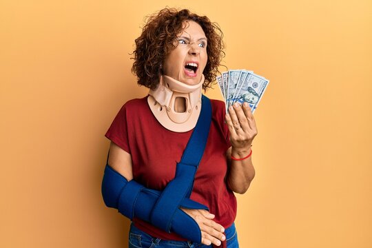 Beautiful middle age mature woman wearing cervical collar and sling holding insurance dollars angry and mad screaming frustrated and furious, shouting with anger. rage and aggressive concept.