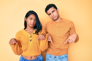 Beautiful latin young couple wearing casual clothes together pointing down looking sad and upset, indicating direction with fingers, unhappy and depressed.