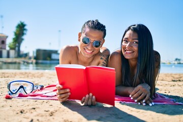 Young latin couple wearing swimwear lying on the sand reading book at the beach.