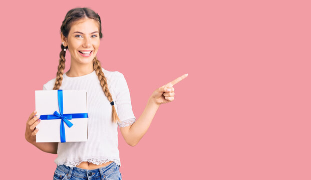 Beautiful caucasian woman with blonde hair holding gift smiling happy pointing with hand and finger to the side