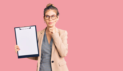 Beautiful caucasian woman with blonde hair holding clipboard with blank space serious face thinking about question with hand on chin, thoughtful about confusing idea