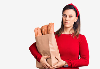 Young beautiful woman holding paper bag with bread thinking attitude and sober expression looking self confident