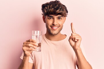 Young hispanic man drinking glass of water smiling with an idea or question pointing finger with happy face, number one